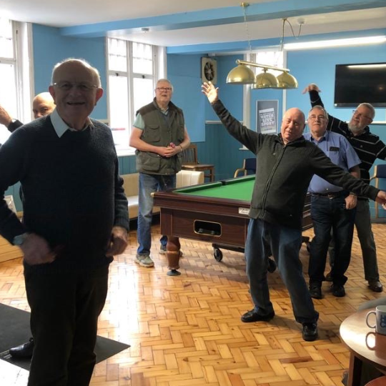Support and Social Connection Men gathered in a bright community room, chatting and laughing—representing the relaxed and welcoming atmosphere of HCCN’s support groups.