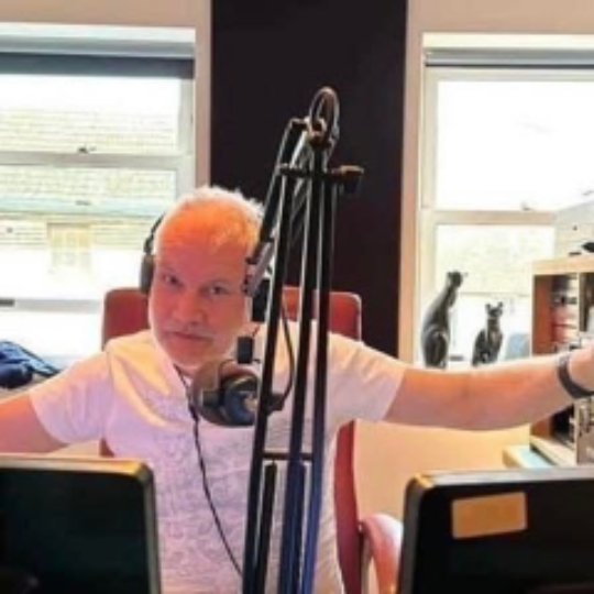 Ste Greenall speaking into a microphone during his breakfast show at Black Cat Radio, supporting HCCN as a community patron.